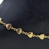 Impon Anklets, a gold-toned chain made of linked flat hearts with three dangling bell charms on one end displayed against a black background