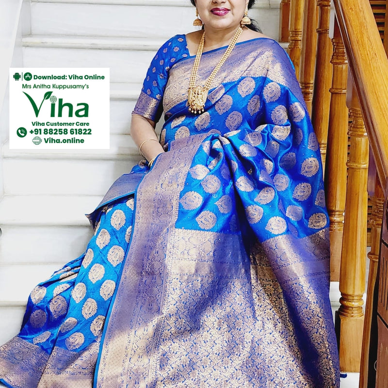Banaras Semi Silk Saree, a woman seated on white stairs wears a blue and gold-toned patterned saree with matching blouse and jewellery