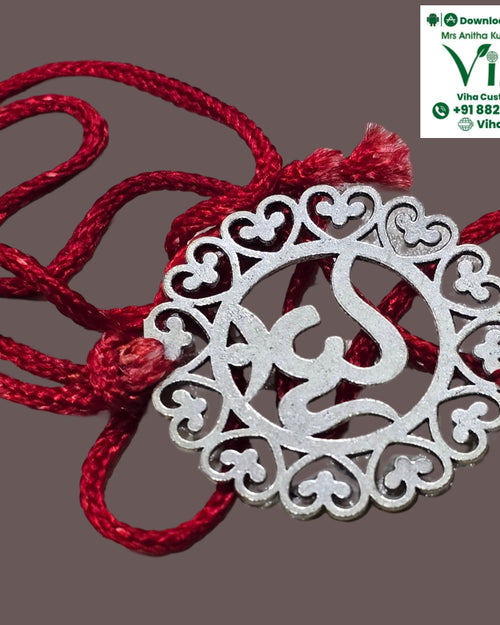 White Metal Rakhi, a silver-toned circular pendant featuring an ornate cut-out symbol is attached to one twisted red string loop