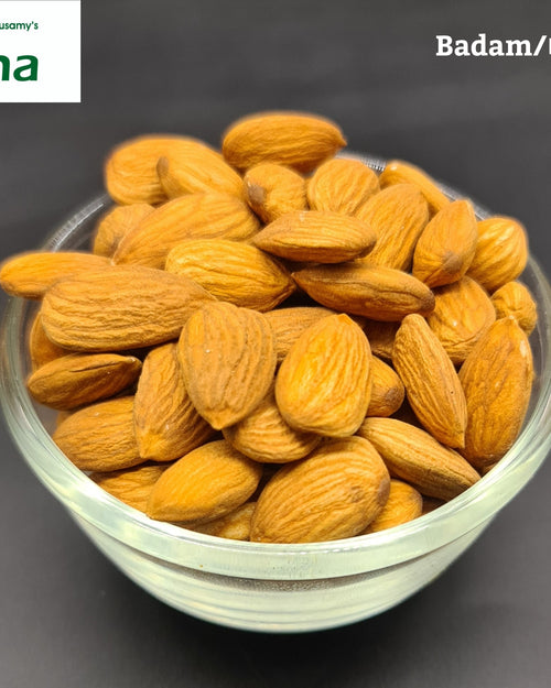 Almond, a clear glass bowl filled with numerous ribbed, brown teardrop-shaped seeds sits against a dark grey background