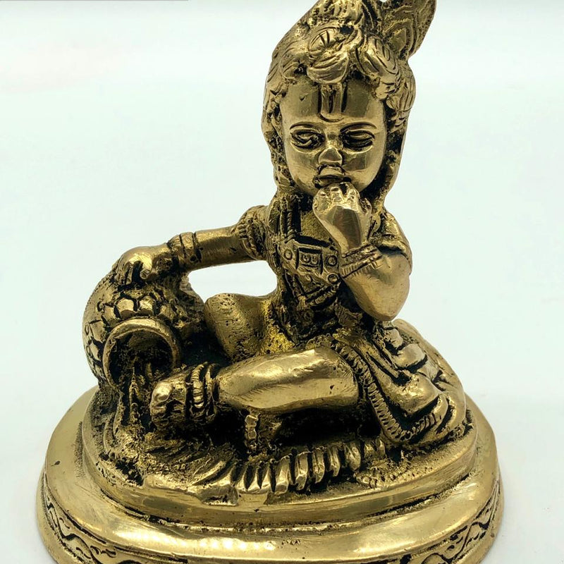 Krishna in brass, child figure seated on a round engraved base with one hand raised to the mouth