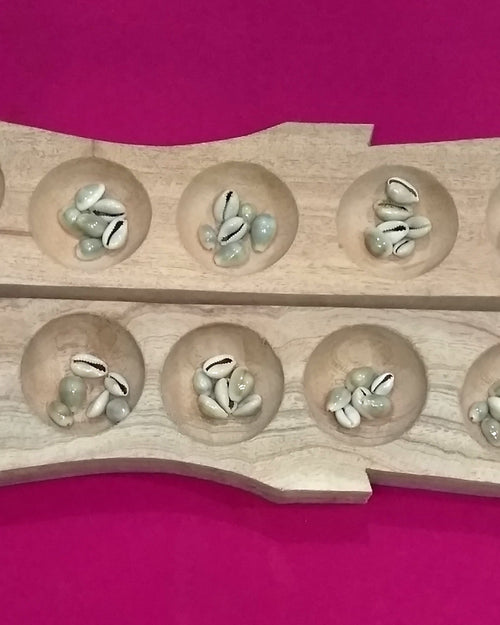 Pallanguzhi With Chozhi, a light brown wooden board with fourteen circular hollows containing small greyish white oval cowrie shells against a pink surface