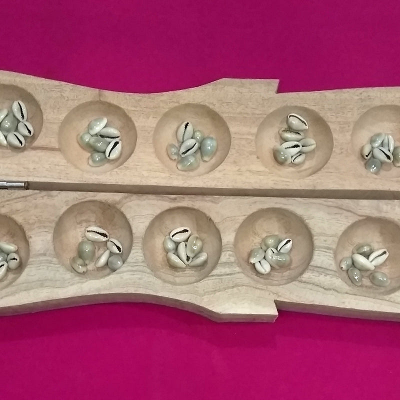 Pallanguzhi With Chozhi, a light brown wooden board with fourteen circular hollows containing small greyish white oval cowrie shells against a pink surface