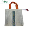 Jute Bag with Zip, off-white woven textured square bag featuring two orange handles and a vertical blue-and-gold-toned patterned ribbon centered on the face