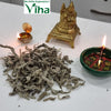 Kubera Mooligai Thiri, a gold-toned figurine, two lit oil lamps, and a pile of greyish dried herbs on a white surface