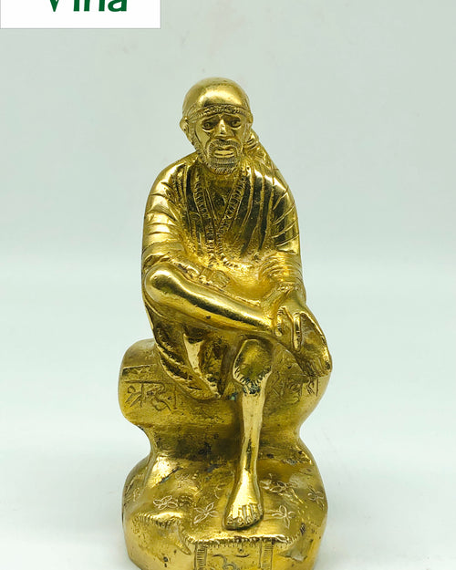 Sai Baba in brass, seated figure leaning forward with a hand on the raised knee, on a rock-shaped pedestal