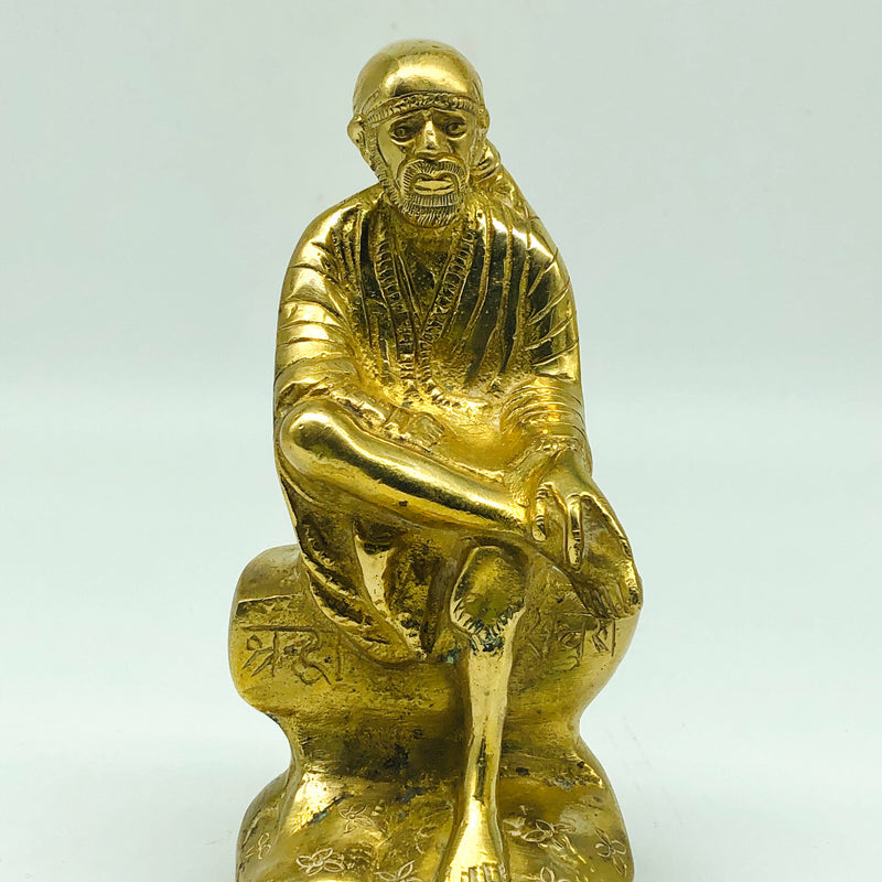 Sai Baba in brass, seated figure leaning forward with a hand on the raised knee, on a rock-shaped pedestal