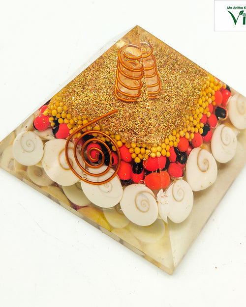 Gomati Chakra/Kundrinmani/Venkadugu Vastu Pyramid, a glossy pyramidal resin object filled with twelve spiral shells, red seeds, and two copper-toned coils